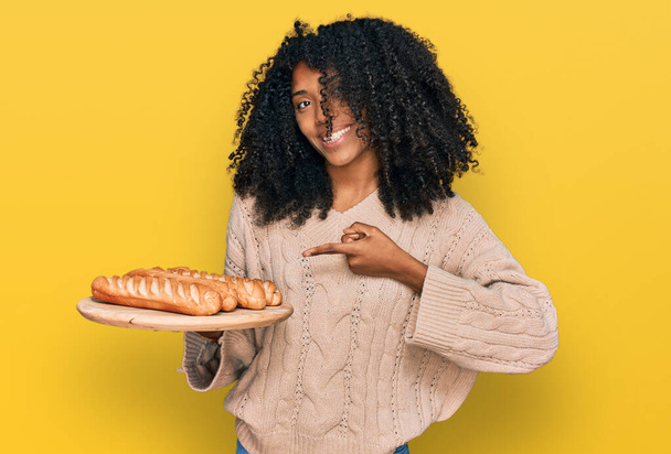 Joven afroamericana americana sosteniendo bandeja con pan sonriendo feliz señalando con la mano y el dedo  - Foto, Imagen
