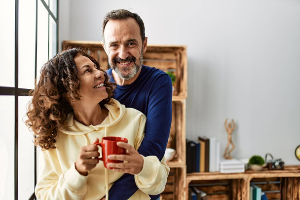 Ein hispanisches Paar mittleren Alters lächelt glücklich und trinkt Kaffee. Zuhause ans Fenster gelehnt. - Foto, Bild