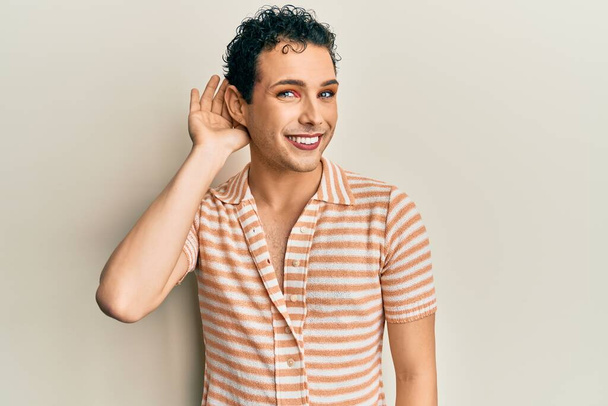 Pohledný muž, který má na sobě make-up, nosí neformální tričko, usmívá se rukou přes ucho a naslouchá drbům a drbům. koncepce hluchoty.  - Fotografie, Obrázek