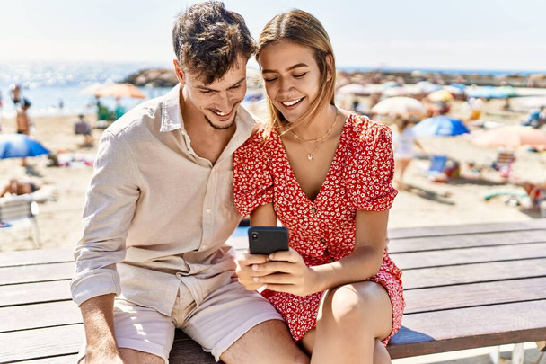 Young hispanic couple on vacation using smartphone sitting on bench at the beach. - Φωτογραφία, εικόνα