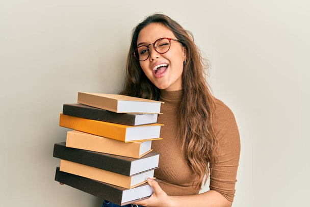 Joven chica hispana sosteniendo un montón de libros guiñando el ojo mirando a la cámara con expresión sexy, cara alegre y feliz.  - Foto, imagen