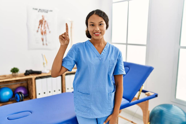 Beautiful hispanic physiotherapist woman at pain recovery clinic showing and pointing up with finger number one while smiling confident and happy.  - Photo, Image