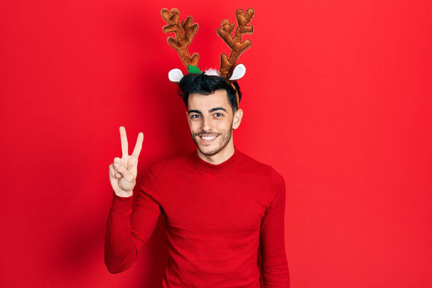 Fiatal spanyol férfi, aranyos karácsonyi rénszarvas szarvakat visel, mutatva és mutatva a kettes számú ujjaival, miközben magabiztosan és boldogan mosolyog..  - Fotó, kép