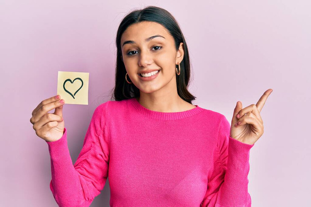 Young hispanic woman holding heart reminder smiling happy pointing with hand and finger to the side  - Photo, Image