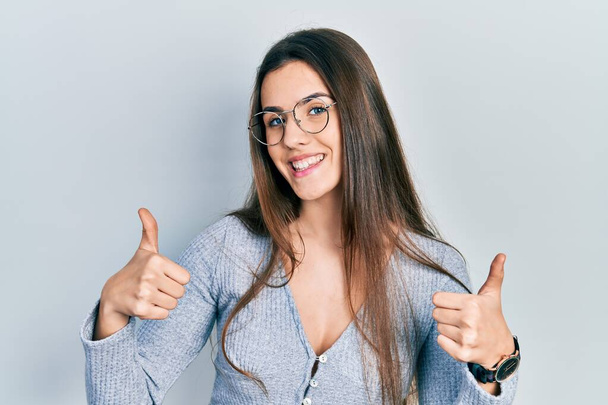若いブルネットのティーンエイジャーは、笑顔と幸せを親指アップ、手で積極的なジェスチャーを行うカジュアルなセーターと眼鏡の成功のサインを着て。陽気な表情と勝者のジェスチャー.  - 写真・画像