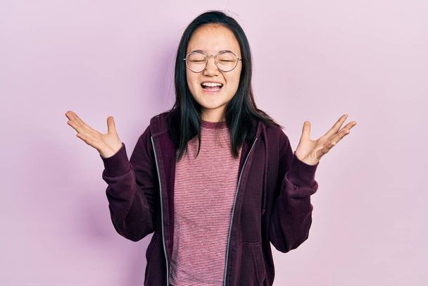 Junges chinesisches Mädchen in lässiger Kleidung und Brille feiert verrückt und verrückt nach Erfolg mit erhobenen Armen und geschlossenen Augen, die aufgeregt schreien. Siegerkonzept  - Foto, Bild