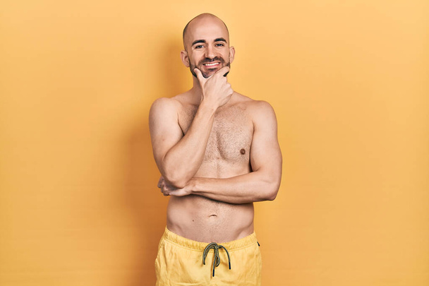 Young bald man wearing swimwear looking confident at the camera smiling with crossed arms and hand raised on chin. thinking positive.  - Fotografie, Obrázek