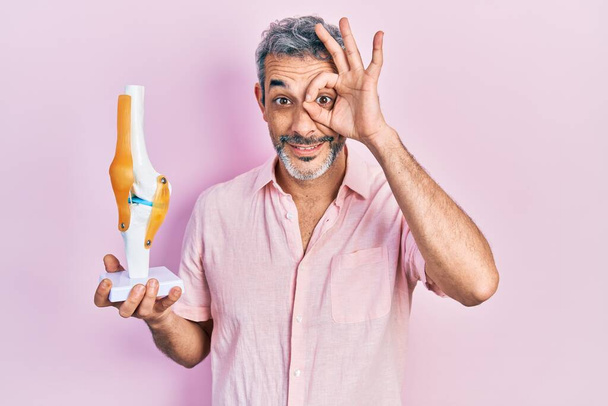 Hombre guapo de mediana edad con pelo gris sosteniendo el modelo anatómico de la articulación de la rodilla sonriendo feliz haciendo signo de ok con la mano en el ojo mirando a través de los dedos  - Foto, imagen