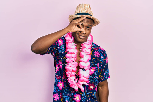 Young black man wearing summer shirt and hawaiian lei smiling happy doing ok sign with hand on eye looking through fingers  - Photo, Image