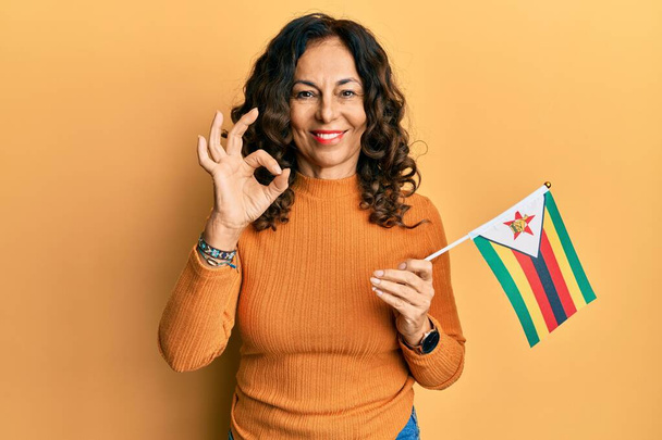 Middelbare leeftijd Spaanse vrouw met zimbabwe vlag doet ok teken met de vingers, glimlachend vriendelijk gebaar uitstekende symbool  - Foto, afbeelding