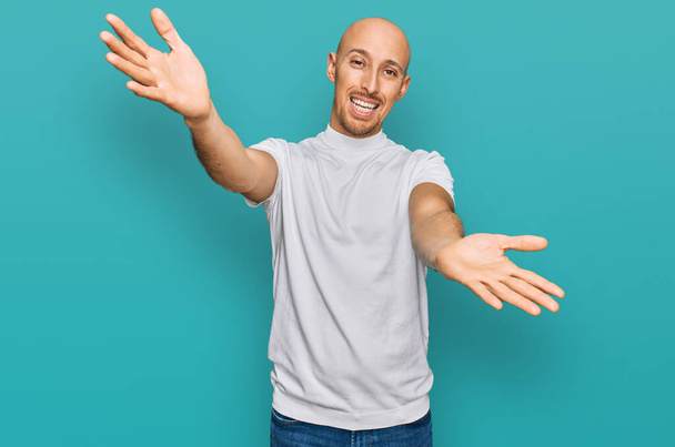 Bald man with beard wearing casual white t shirt looking at the camera smiling with open arms for hug. cheerful expression embracing happiness.  - Photo, Image