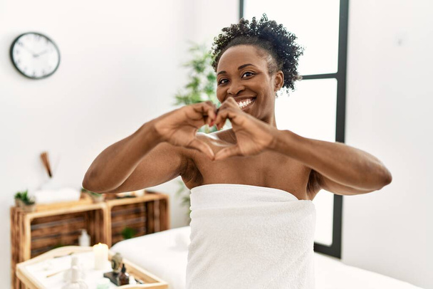 Mujer afroamericana joven con toalla de pie en el centro de belleza sonriendo en el amor mostrando el símbolo del corazón y la forma con las manos. concepto romántico.  - Foto, imagen