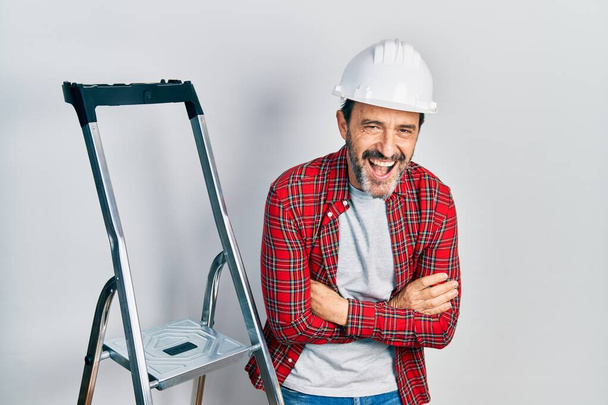Obrero hispano de mediana edad con los brazos cruzados gesto usando escalera sonriendo y riendo en voz alta porque divertido chiste loco.  - Foto, imagen