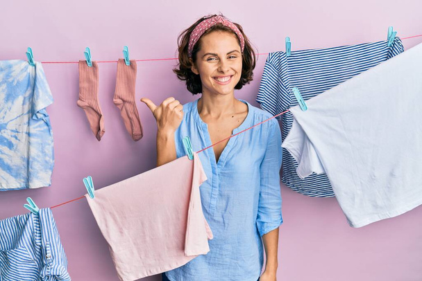 Young brunette woman doing laundry around string hangs smiling with happy face looking and pointing to the side with thumb up.  - Foto, Imagem