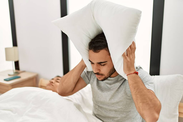 Мужчина покрывает уши подушкой, раздраженной за храп, лежащий в постели дома. - Фото, изображение