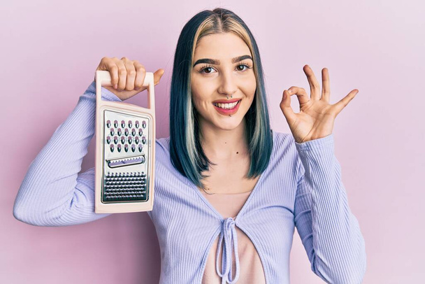 Young modern girl holding grater doing ok sign with fingers, smiling friendly gesturing excellent symbol  - Zdjęcie, obraz