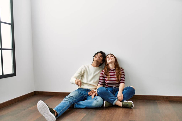 Jovem casal hispânico olhando para o teto sentado no chão em casa nova vazia. - Foto, Imagem