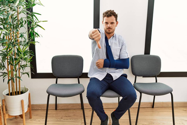 Handsome young man sitting at doctor waiting room with arm injury looking unhappy and angry showing rejection and negative with thumbs down gesture. bad expression.  - Photo, Image
