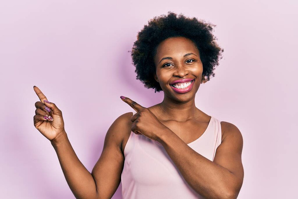 Young african american woman wearing casual sleeveless t shirt smiling and looking at the camera pointing with two hands and fingers to the side.  - Photo, Image