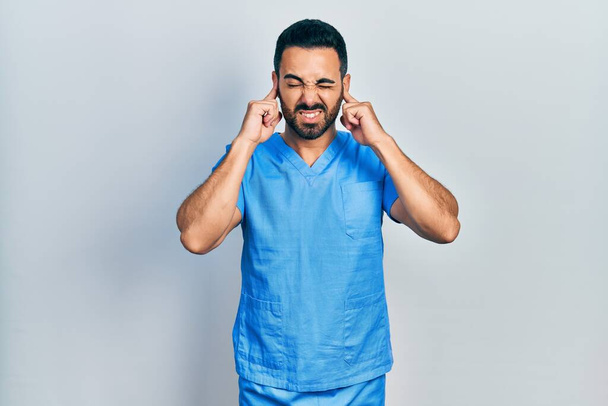 Hombre hispano guapo con barba vistiendo uniforme de enfermero azul cubriendo oídos con dedos con expresión molesta por el ruido de la música alta. concepto de sordo.  - Foto, imagen
