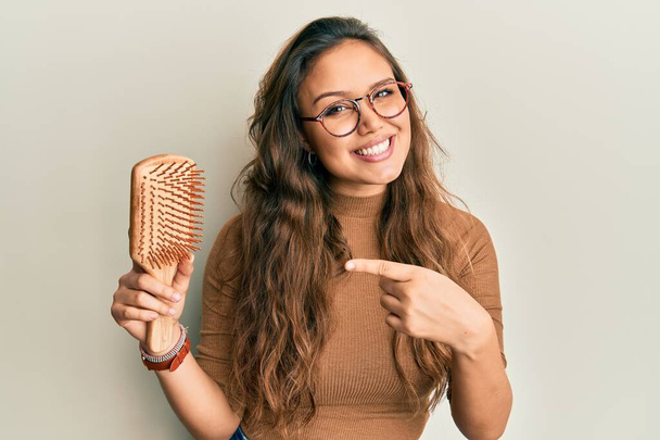 Young hispanic girl holding hair comb smiling happy pointing with hand and finger  - 写真・画像