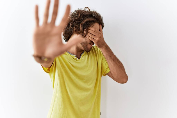 Молодий іспаномовний чоловік стоїть над ізольованим тлом, закриваючи очі руками і роблячи стоп жест з сумним і страховим виразом. збентежена і негативна концепція
.  - Фото, зображення