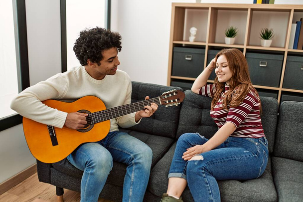 Pareja joven sonriendo feliz tocando la guitarra clásica sentada en el sofá en casa. - Foto, imagen