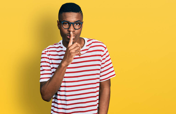 A fiatal afro-amerikai férfi alkalmi pólót és szemüveget visel, és azt kéri, hogy maradjon csendben ujjal a száján. csend és titkos koncepció.  - Fotó, kép