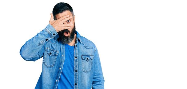 Homme hispanique avec barbe veste en denim décontracté jetant un coup d'oeil dans le visage de couverture de choc et les yeux avec la main, regardant à travers les doigts peur  - Photo, image