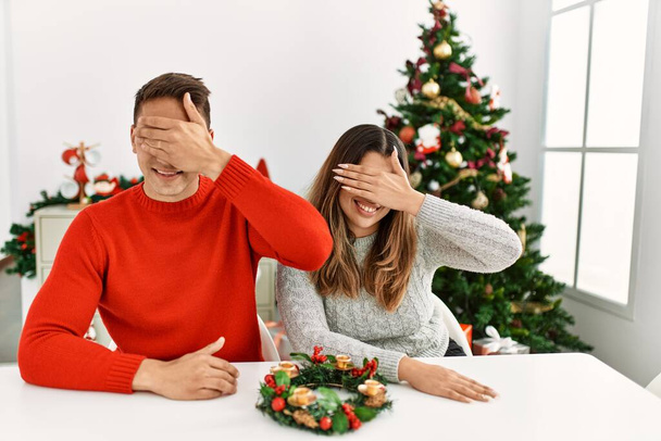 Ein junges hispanisches Paar, das an Weihnachten am Tisch sitzt, lächelt und lacht und die Augen zur Überraschung mit der Hand auf dem Gesicht verdeckt. Blindes Konzept.  - Foto, Bild