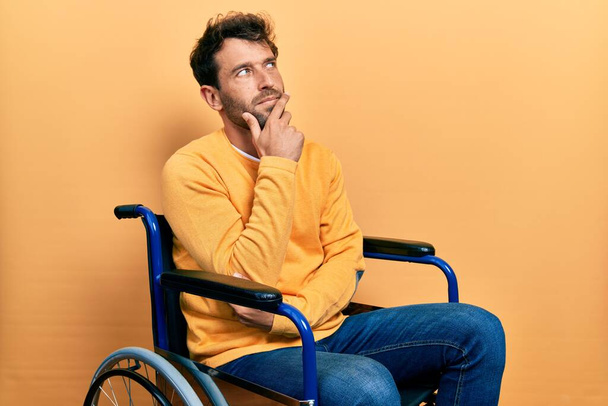 Όμορφος άντρας με γενειάδα κάθεται σε αναπηρική καρέκλα με το χέρι στο πηγούνι σκέφτεται την ερώτηση, σκεπτική έκφραση. Χαμογελώντας με ευγενικό πρόσωπο. έννοια της αμφιβολίας.  - Φωτογραφία, εικόνα