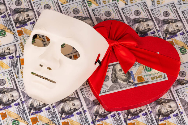 Een $100 biljet ligt op een hartvormige geschenkdoos naast een plastic wit masker tegen een achtergrond van $100 biljetten - Foto, afbeelding