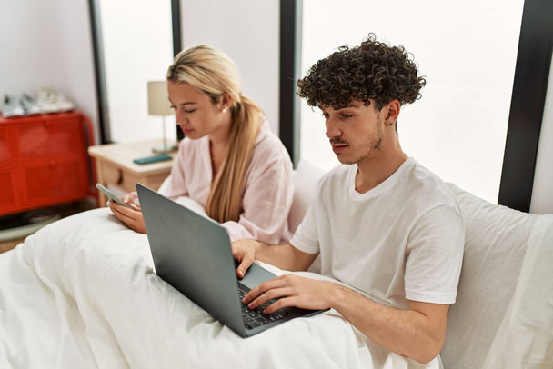 Young beautiful couple using laptop and smartphone sitting on the bed at home. - Фото, зображення
