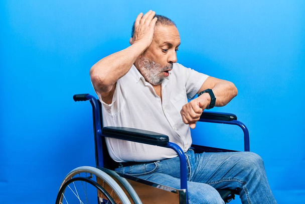 ひげを生やしたハンサムな老人が車椅子に座って時計の時間を見て心配し、遅くなるのを恐れて  - 写真・画像