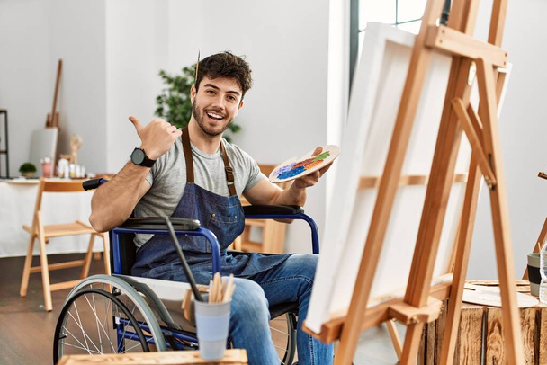 Sanat stüdyosundaki tekerlekli sandalyede oturan genç adam, arka tarafı işaret ediyor, el ve baş parmakları havada, kendinden emin gülümsüyor.  - Fotoğraf, Görsel