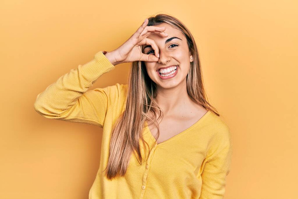 Hermosa mujer hispana vistiendo suéter amarillo casual haciendo buen gesto con la mano sonriente, ojos mirando a través de los dedos con la cara feliz.  - Foto, Imagen
