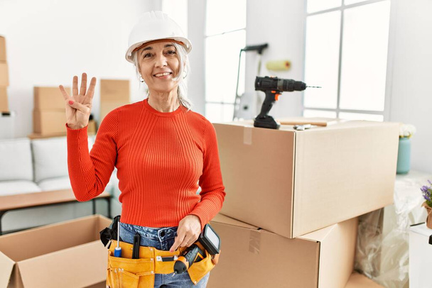 Siwowłosa kobieta w średnim wieku w twardym kapeluszu stojąca w nowym domu, pokazująca i wskazująca palcami numer cztery, uśmiechnięta pewna siebie i szczęśliwa..  - Zdjęcie, obraz