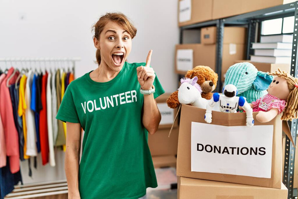 Прекрасная кавказская женщина в футболке волонтера на пожертвованиях стоит, показывая пальцем вверх с успешной идеей. Взволнован и счастлив. номер один.  - Фото, изображение