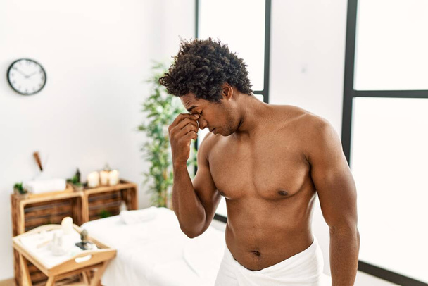 Młody Afroamerykanin bez koszulki noszący ręcznik stojący w centrum piękności zmęczony pocieraniem nosa i oczu odczuwający zmęczenie i ból głowy. koncepcja stresu i frustracji.  - Zdjęcie, obraz