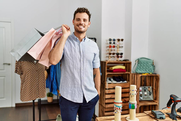 Young hispanic customer man smiling happy holding shopping bags at clothing store. - Фото, изображение