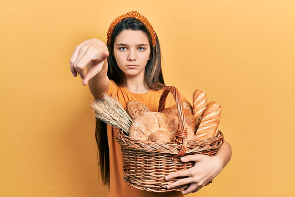 Młoda brunetka dziewczyna trzyma wiklinowy kosz z chlebem wskazując palcem na aparat i do Ciebie, pewny gest patrząc poważnie  - Zdjęcie, obraz