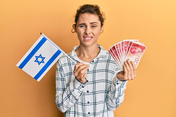 Mladá brunetka držící izraelskou vlajku a šekely bankovky, bezradný a zmatený výraz. pochybný koncept.  - Fotografie, Obrázek