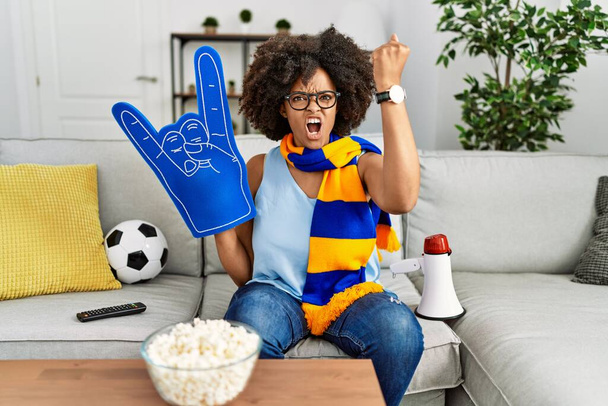 Mujer afroamericana con pelo afro vistiendo bufanda de equipo animando juego sosteniendo megáfono molesto y frustrado gritando con ira, gritando loco de ira y la mano levantada  - Foto, imagen