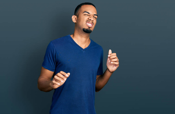 Genç Afro-Amerikalı bir adam günlük tişört giyiyor, tiksindirici bir ifade takınıyor ve tiksindirici bir yüz ifadesi takınıyor. Eller havada.  - Fotoğraf, Görsel