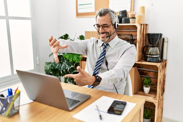 Latynoski biznesmen w średnim wieku, pracujący w biurze, noszący słuchawki operatora, uśmiechający się, robiący ramę rękami i palcami ze szczęśliwą twarzą. koncepcja kreatywności i fotografii.  - Zdjęcie, obraz