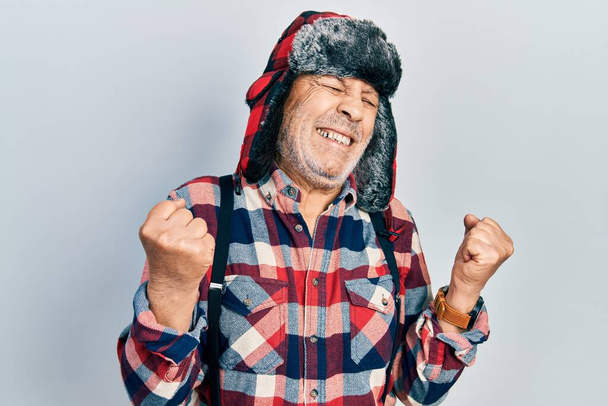 Schöner älterer Mann mit Wintermütze und Ohrenklappen sehr glücklich und aufgeregt dabei Siegergeste mit erhobenen Armen, lächelnd und schreiend nach Erfolg. Festkonzept.  - Foto, Bild