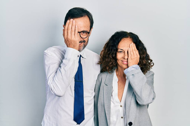 Paar mittleren Alters aus hispanischen Frauen und Männern in Business-Office-Uniform, die ein Auge mit der Hand bedecken, selbstbewusstes Lächeln im Gesicht und überraschende Emotionen.  - Foto, Bild