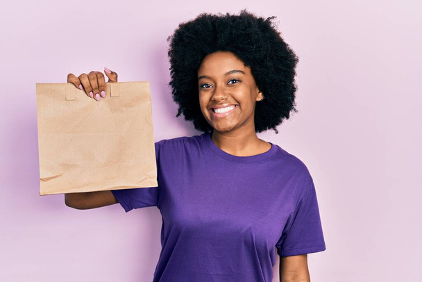 Fiatal afro-amerikai nő gazdaság elvenni papírzacskó néz pozitív és boldog álló és mosolygó magabiztos mosollyal mutatja fogak  - Fotó, kép