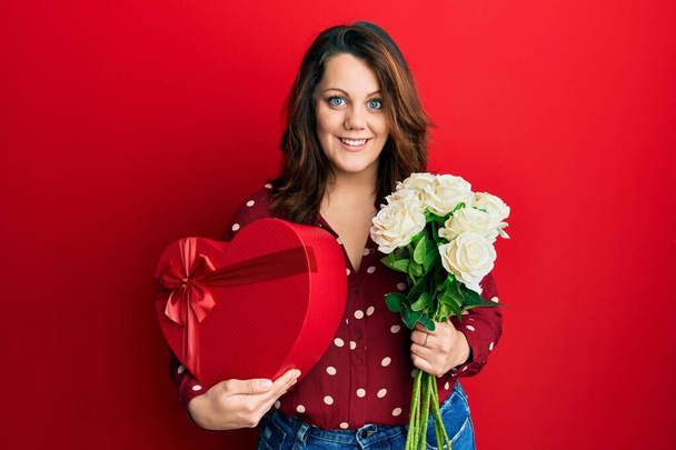 Mujer caucásica joven sosteniendo regalo de San Valentín y flores sonriendo con una sonrisa feliz y fresca en la cara. mostrando dientes.  - Foto, imagen