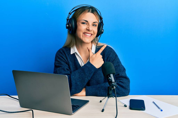 młoda kaukaska kobieta pracuje w radio studio uśmiechając wesoły wskazując ręką i palcem w górę do boku  - Zdjęcie, obraz